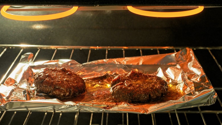 Two burger patties under a broiler on a baking sheet lined with foil