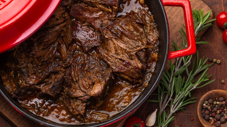 Braised beef in Dutch oven next to rosemary and other seasonings