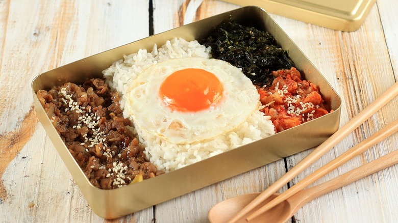 Dosirak in metal tin with wood spoon and chopsticks nearby on wood table