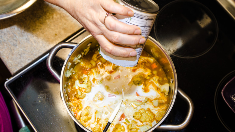 Hand pouring contents of a canned good into a pot with stew or soup on the stovetop.