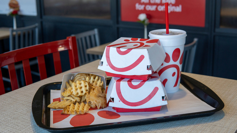 A Chick-fil-A meal sits on a tabletop tray in a Chick-fil-A restaurant: a medium order of waffle fries, two cardboard clamshell boxes, and a medium soft drink.