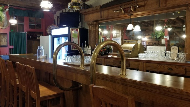 The bar area at The Log Inn is dominated by dark wood, with brass fixtures and pink-painted walls. Glassware sits on the bar and tall barstools are slid beneath the bar.