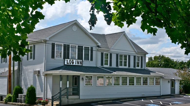 Indiana's oldest restaurant, The Log Inn, is a two-story off-white building with shuttered windows and a wide front. There is a covered porch and leafy trees frame the picture.