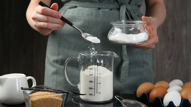 Closeup person scooping baking powder into measuring cup alongside eggs, sugar, and vanilla.