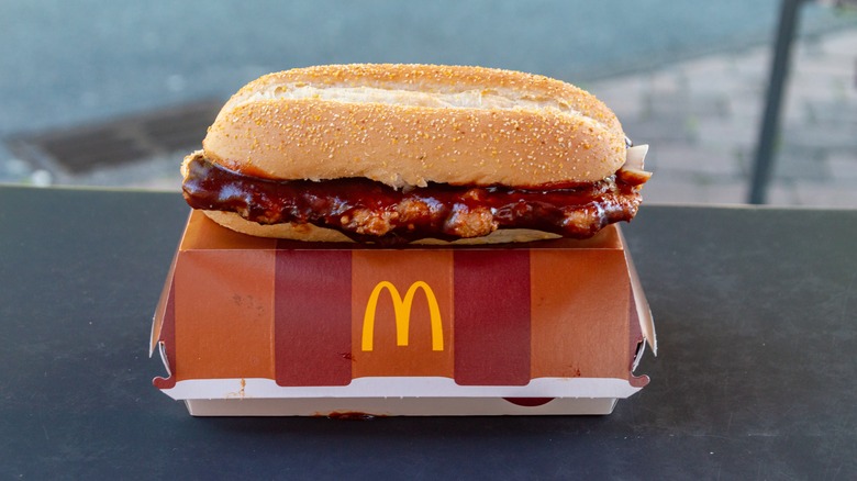 A saucy McRib sandwich sits atop its orange-striped clamshell box in a black surface. Behind it, a window reveals a blurry view of a nondescript street.