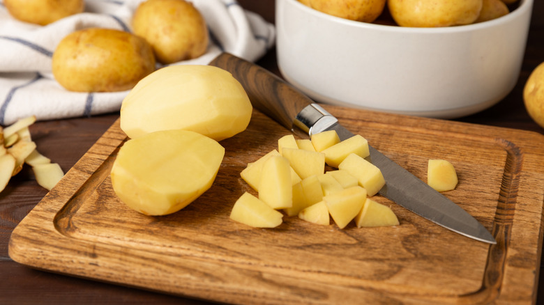 Cubed raw potato on a wooden chopping board, next to a knife and two peeled, raw potatoes