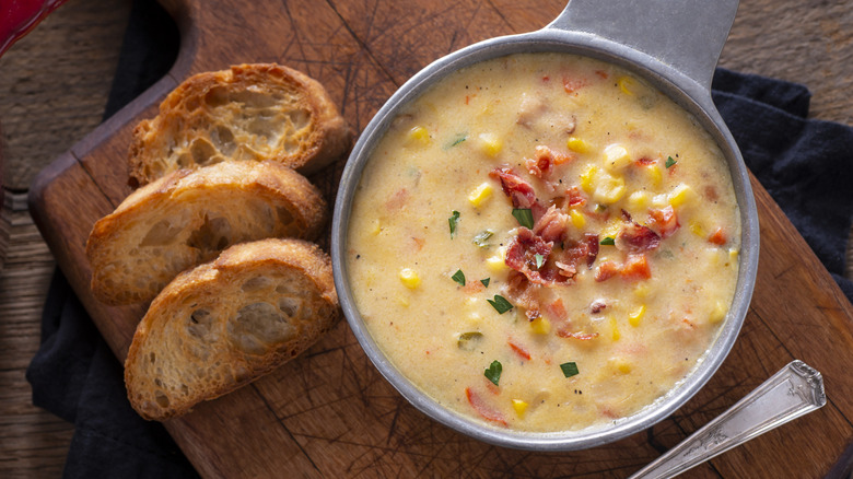 Bowl of corn chowder with bacon bits and spices with three pieces of toasted bread on a wooden board.