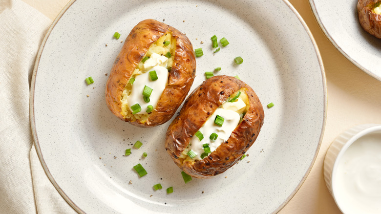 Two baked potatoes sit on a flecked white plate. The potatoes are topped with a generous dollop of sour cream and sprinkled with cut green onions, which are also scattered on the plate.