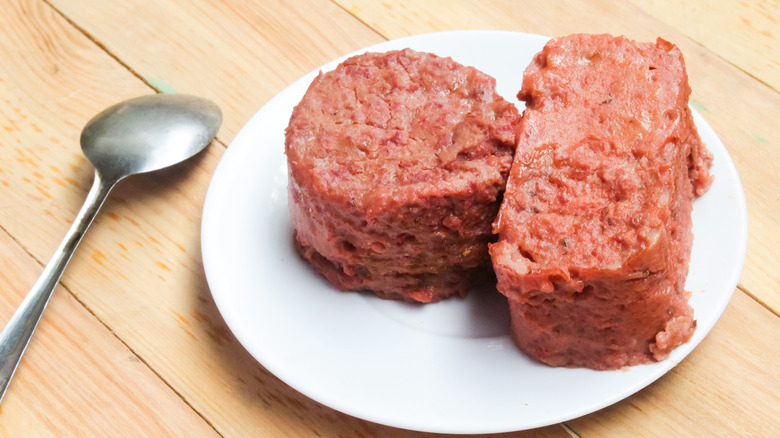 Canned corned beef sits, holding the shape of its container, on a white plate atop a wooden surface, with a silver spoon by its side.