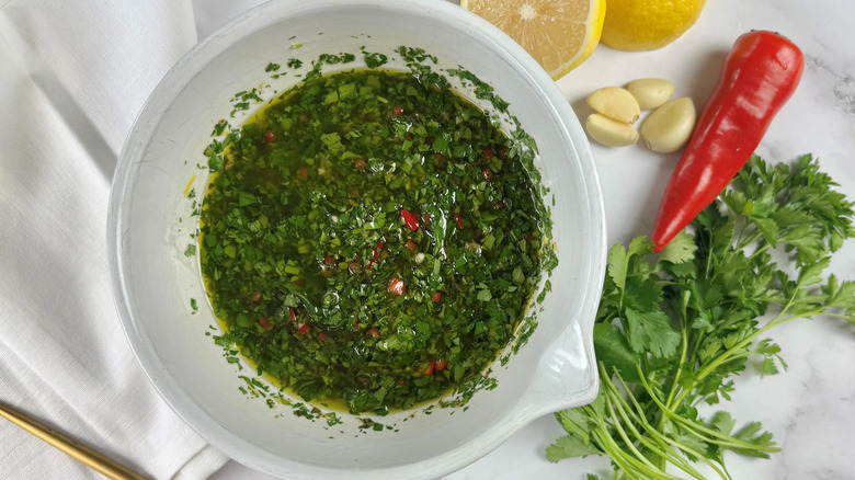 Bowl of chimichurri next to herbs, garlic, chile, and lemon