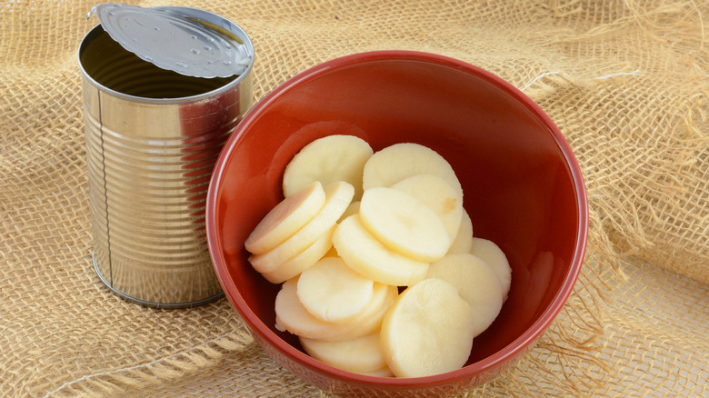 Sliced canned potatoes in a red bowl with opened can on textured background
