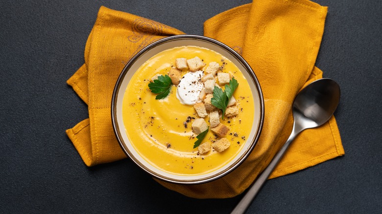 bowl of pumpkin soup with croutons and herbs, atop yellow cloth napkin with spoon to the side.