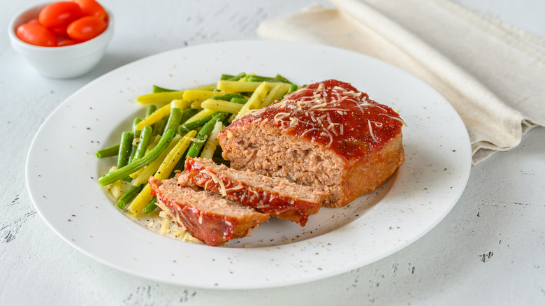 Homemade ground meat meatloaf with ketchup and green beans on white plate.