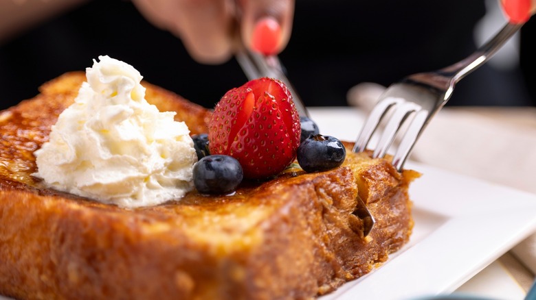 Person cutting into French toast with berries on top using knife and fork