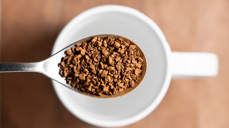 A silver spoonful of instant coffee crystals is raised above an empty white mug on a beige background.