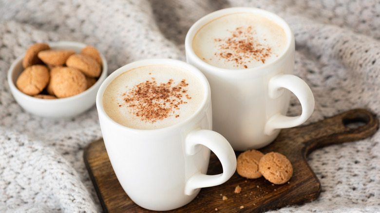 Two cups of coffee in white mugs with frothed milk and a dash of cinnamon sit atop a mini bread board on a knitted blanket with some cookies.