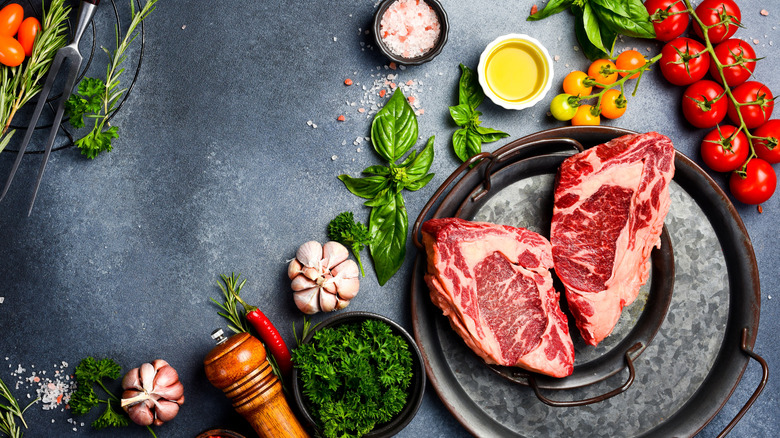 two raw ribeye steaks on a plate surrounded by herbs, seasonings, and tomatoes