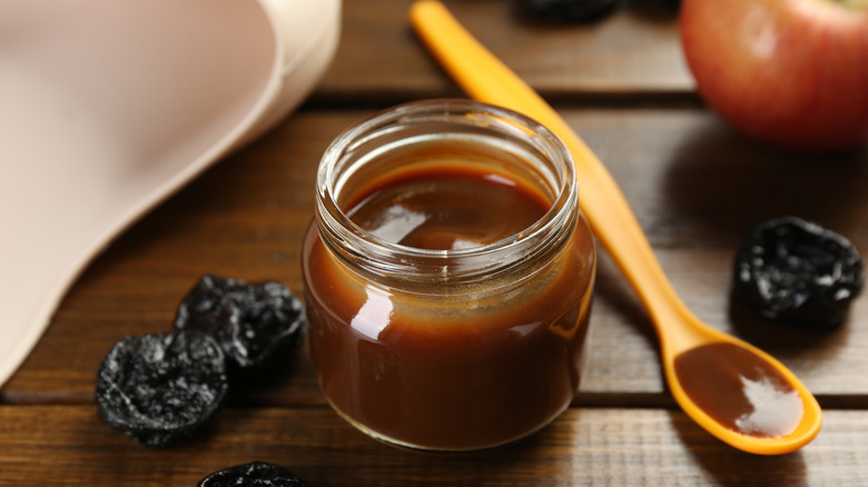 Pureed prunes baby food in jar and prunes on wooden table with a yellow spoon.