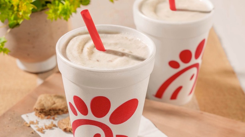 Two Chick-fil-A milkshakes with straws resting beside a snack and a plant