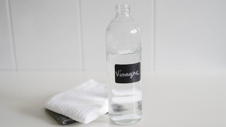 A glass bottle labeled "vinagre" against white tile backdrop with white towel.