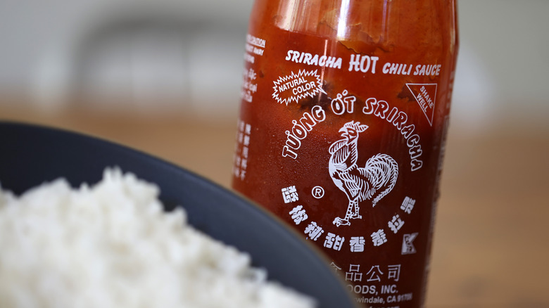 Bottle of Huy Fong sriracha sauce next to blurred bowl of rice.