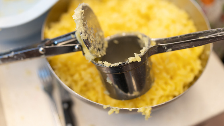 An open potato ricer above pot of mashed yellow potatoes