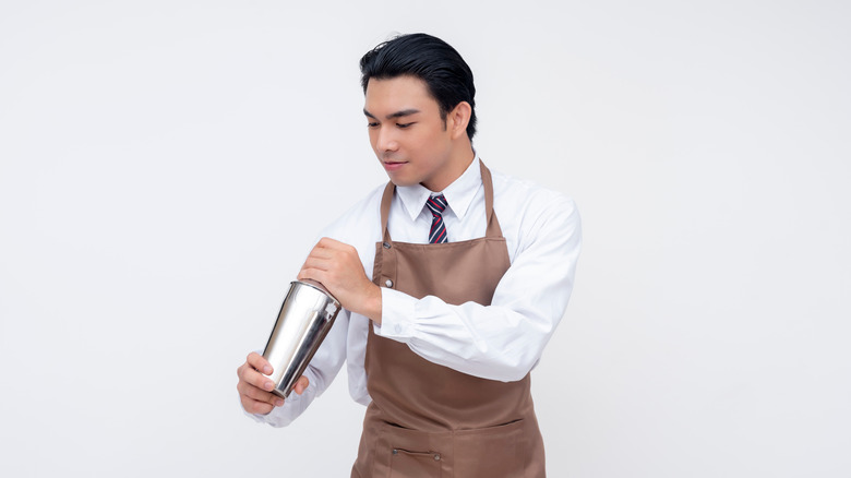 A bartender in a brown apron holds a cocktail shaker