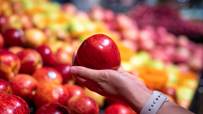 Hand holding supermarket apple with a glossy wax coating against blurred apple background.