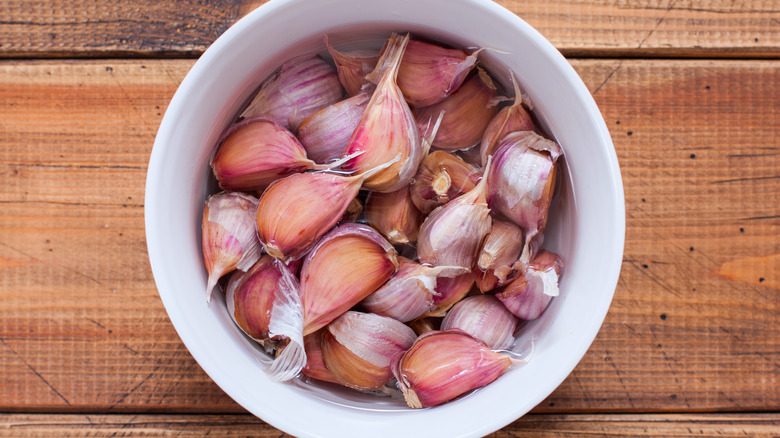 Whole garlic cloves with peels in a white bowl of water. 