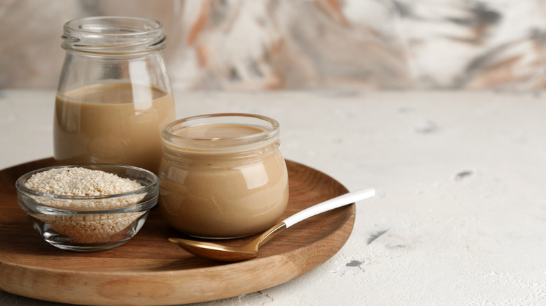 Jars of tahini and ramekin of sesame seeds on wooden dish