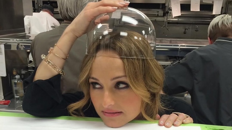 Giada De Laurentiis resting her chin on a table and holding a glass cloche on her head