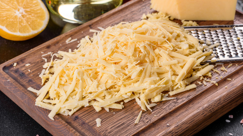 Fresh hard cheese grated on a large grater on a wooden cutting board on a dark background with a lemon wedge in the corner..