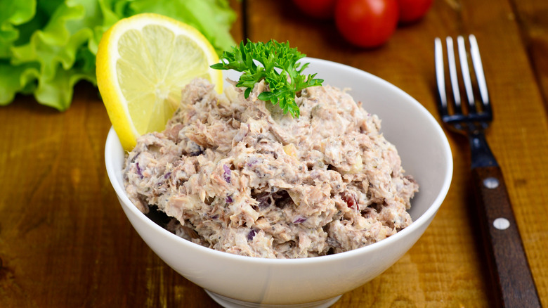 Tuna salad in a white bowl with a slice of lemon and parsley on a wooden table with a fork.