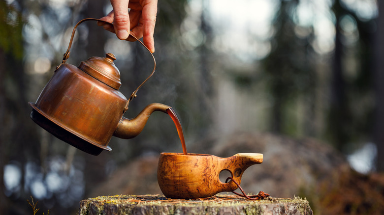 Person pouring coffee from a copper kettle into a wooden mug