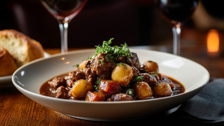 Beef stew in white bowl with wine glasses in background