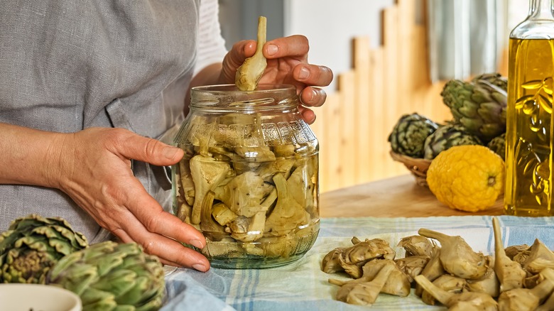 A person's hands handle canned artichokes in a big glass jar.