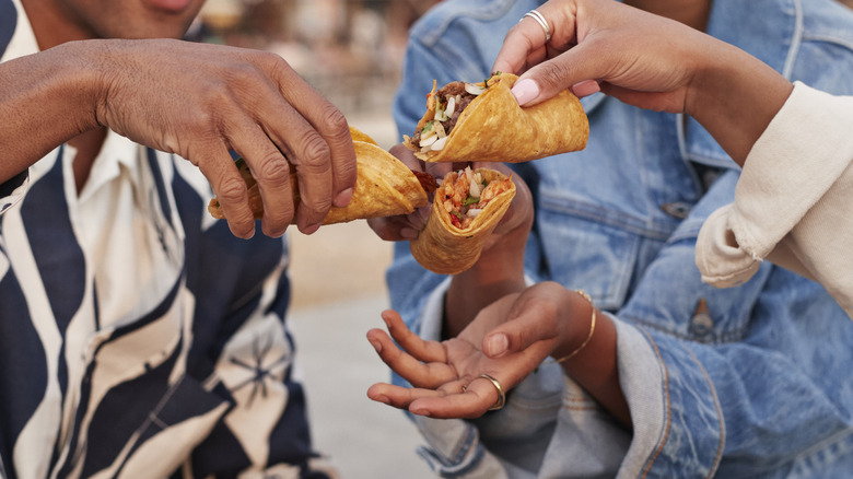 Friends gather to share tacos, which are rolled in corn tortillas and help in three outstretched hands.