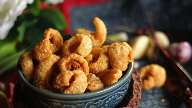 Crispy, golden chicharrons are piles high in a blue, flowered bowl atop a woven charger with a blurred, busy background.