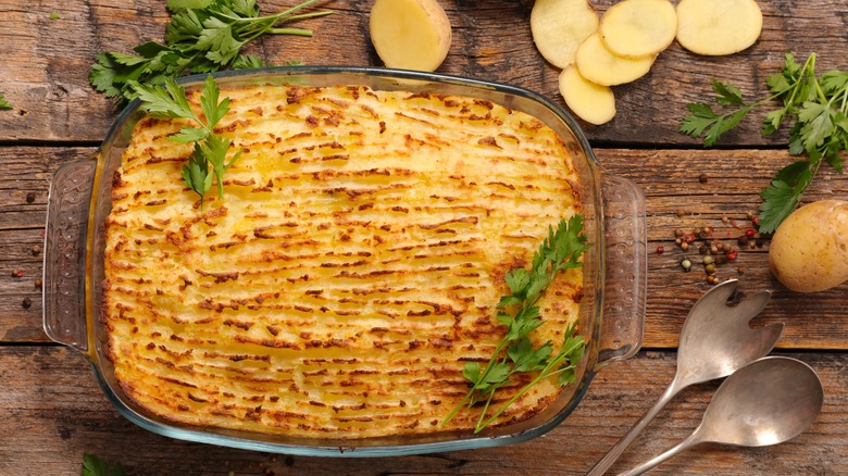 A rustic shepherd's pie with a golden top of furrowed potatoes sits on a rustic wooden surface with sliced potatoes and parsley around the baking dish.