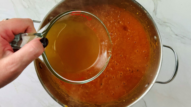 hand pouring stock into pureed tomatoes