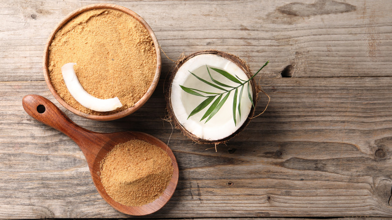 Coconut sugar in bowl, spoon, palm leaves and fruit on wooden table.