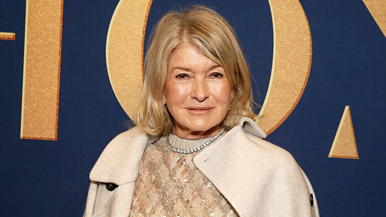 Martha Stewart wears a champagne-colored coat and a diamond necklace in front of a navy blue and gold background on a red carpet.