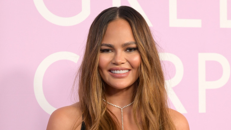 Chrissy Teigen in front of a pink and white sign, smiling