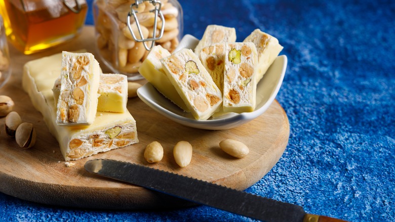 Nougat candy cut into pieces, showing pistachios and almonds, on wooden cutting board with serrated knife in foreground.