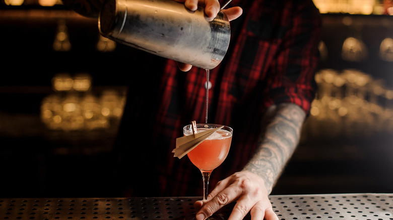 bartender pouring a paper plane cocktail from a strainer into a coupe glass with a paper plane garnish