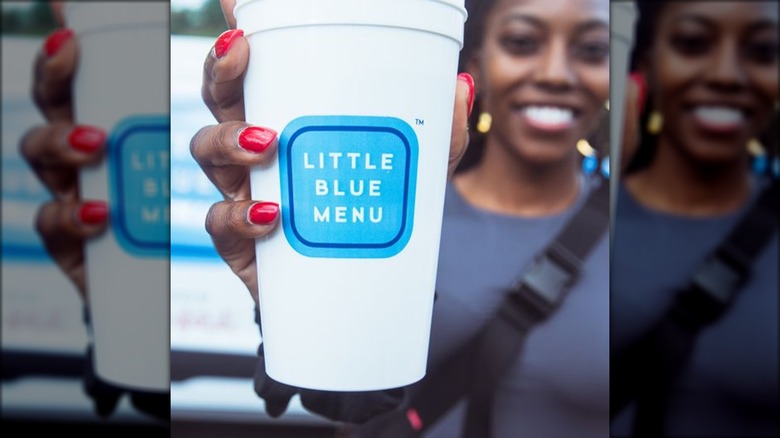 A smiling woman with red fingernails holds a white and blue Little Blue Menu disposable cup up to the camera.