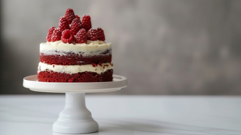 Red velvet cake with cream cheese frosting and raspberries on white cake stand.