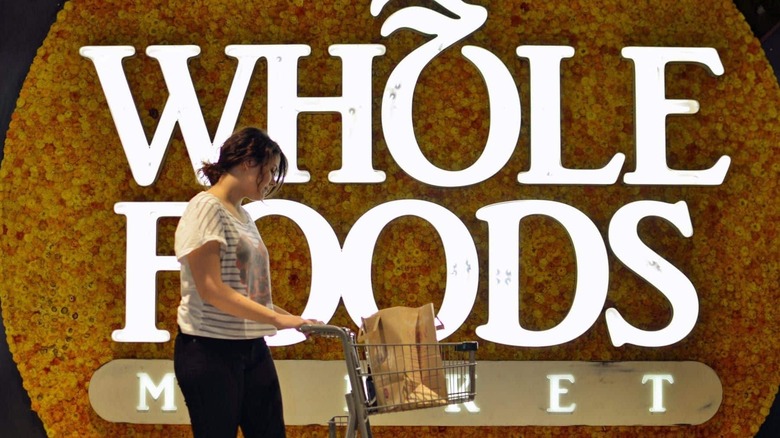 A woman with a mini shopping cart stands in front of the Whole Foods Market logo on a wall