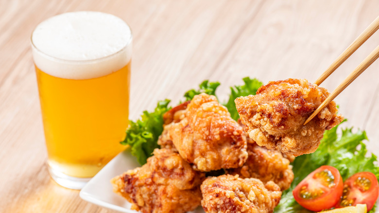 small piece of fried chicken being held by chopsticks with a salad and beer in the background