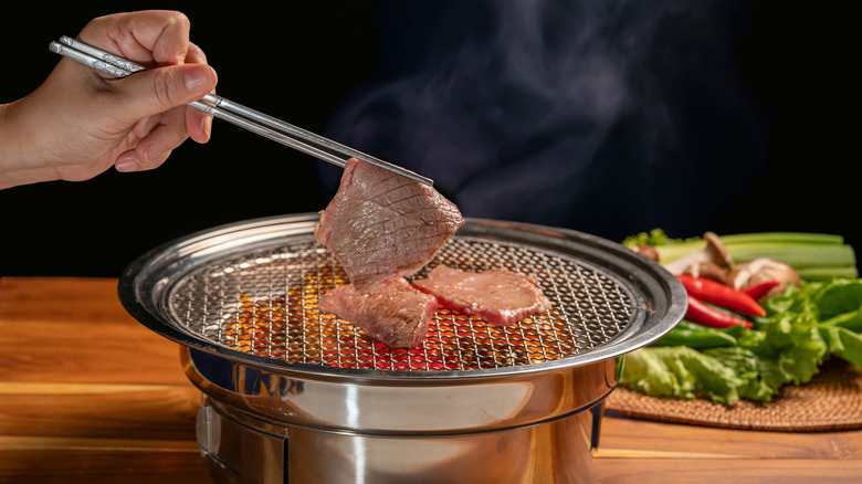 A hand using chopsticks to pick up a thin piece of meat from a tabletop grill, vegetables are in the background.